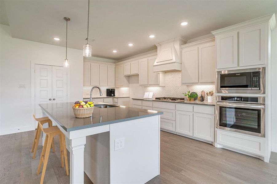 Kitchen featuring light hardwood / wood-style floors, tasteful backsplash, an island with sink, premium range hood, and appliances with stainless steel finishes