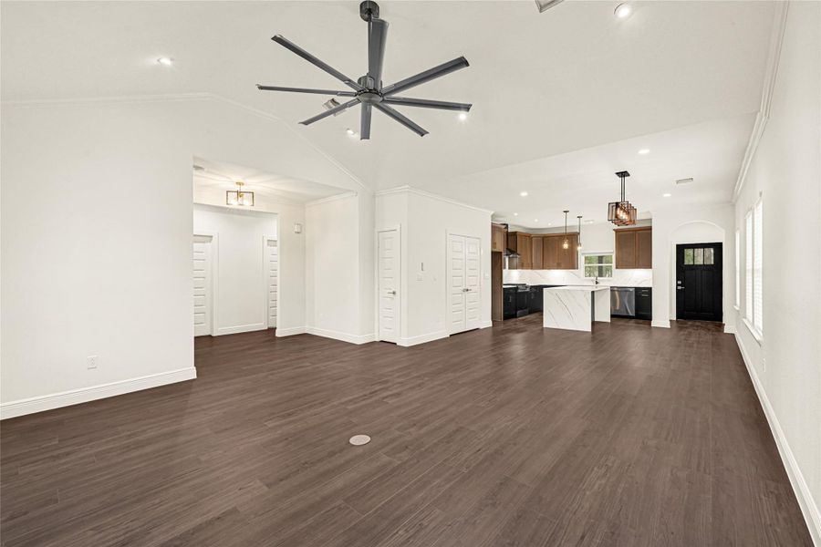 This is a spacious, open-concept living area with modern fixtures and dark wood flooring. It features a large ceiling fan, recessed lighting, and a view into a sleek kitchen with an island and pendant lights. A black entry door and multiple windows provide natural light. This is a spacious, open-concept living area with modern fixtures and dark wood flooring. It features a large ceiling fan, recessed lighting, and a view into a sleek kitchen with an island and pendant lights. A black entry door and multiple windows provide natural light.