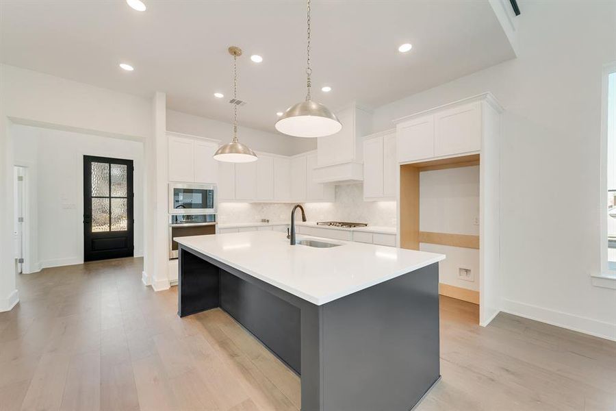 Kitchen featuring white cabinets, backsplash, decorative light fixtures, a center island with sink, and stainless steel appliances Kitchen featuring white cabinets, backsplash, decorative light fixtures, a center island with sink, and stainless steel appliances