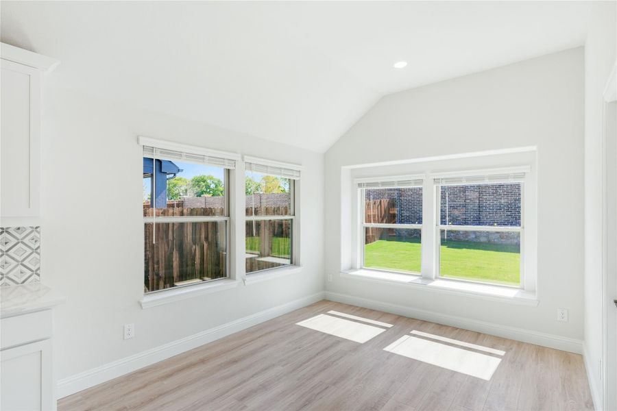 Empty room with vaulted ceiling, light wood floors, and large windows overlooking a fenced backyard