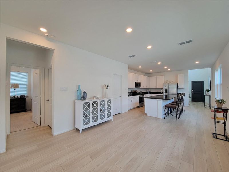 Bright, open-concept kitchen and living area with light wood flooring. The kitchen features modern white cabinetry, stainless steel appliances, and a central island with seating. A doorway leads to a cozy bedroom, and the space is well-lit with recessed lighting.