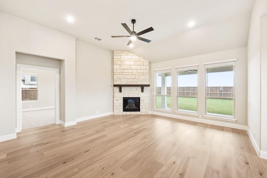 Open living room with stone fireplace, hardwood floors, ceiling fan, and large windows overlooking backyard
