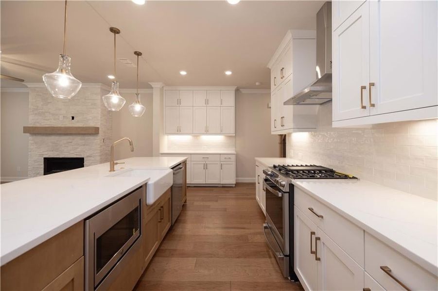 Kitchen with appliances with stainless steel finishes, wall chimney exhaust hood, white cabinetry, hanging light fixtures, and ornamental molding