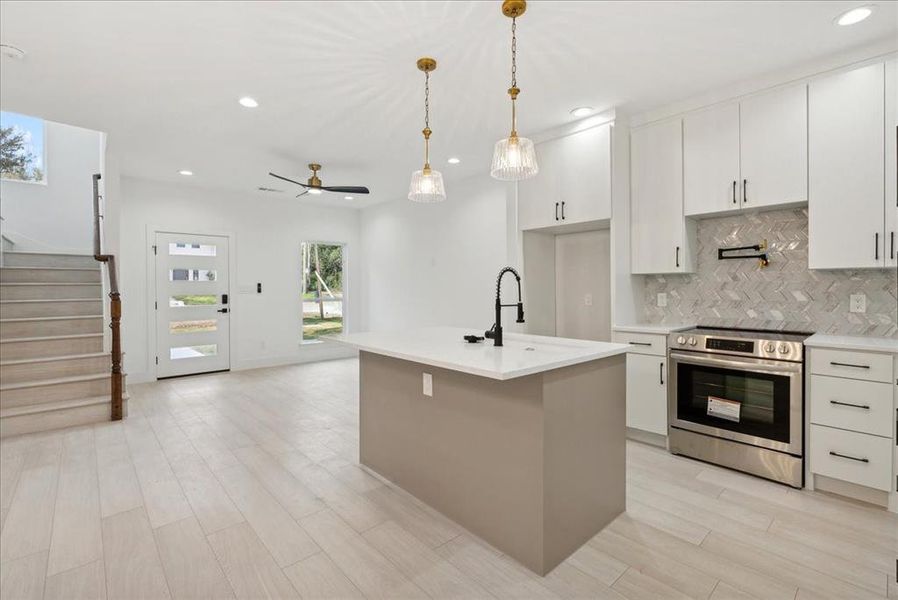 Kitchen featuring stainless steel electric stove, a center island with sink, sink, decorative light fixtures, and white cabinetry