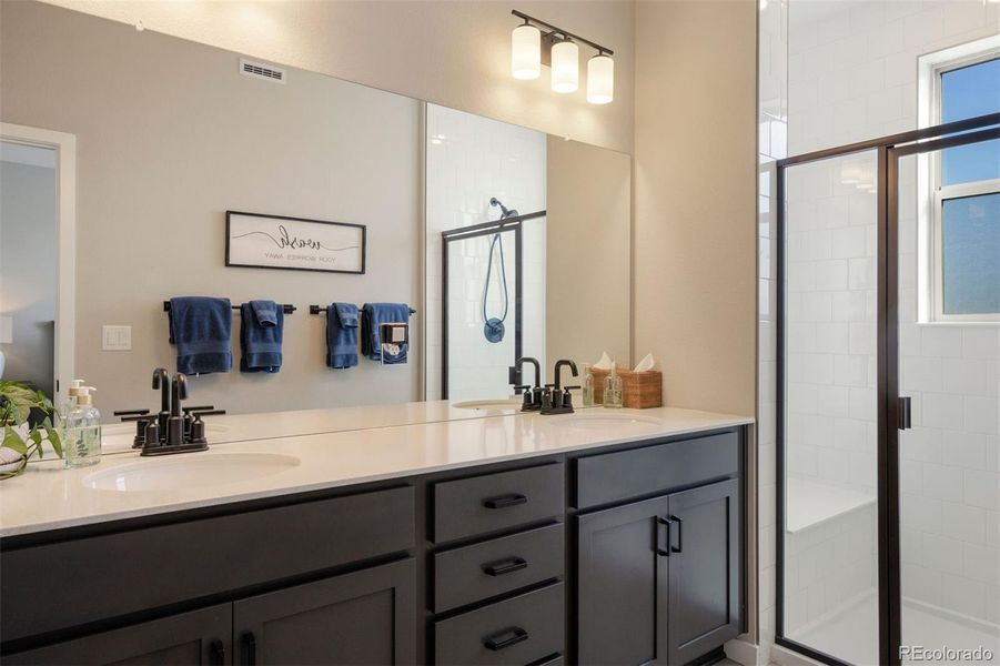 Spacious primary bathroom with dual vanities, a large mirror, abundant cabinets with soft-close functionality and a glass faced shower with subway tile surround.