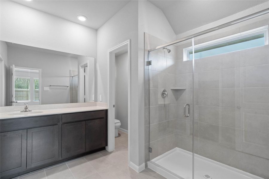A bright and functional primary bath offering a long stained cabinet for extra storage, light countertop, a large mirror, and a modern glass-enclosed shower, creating a serene and efficient space.