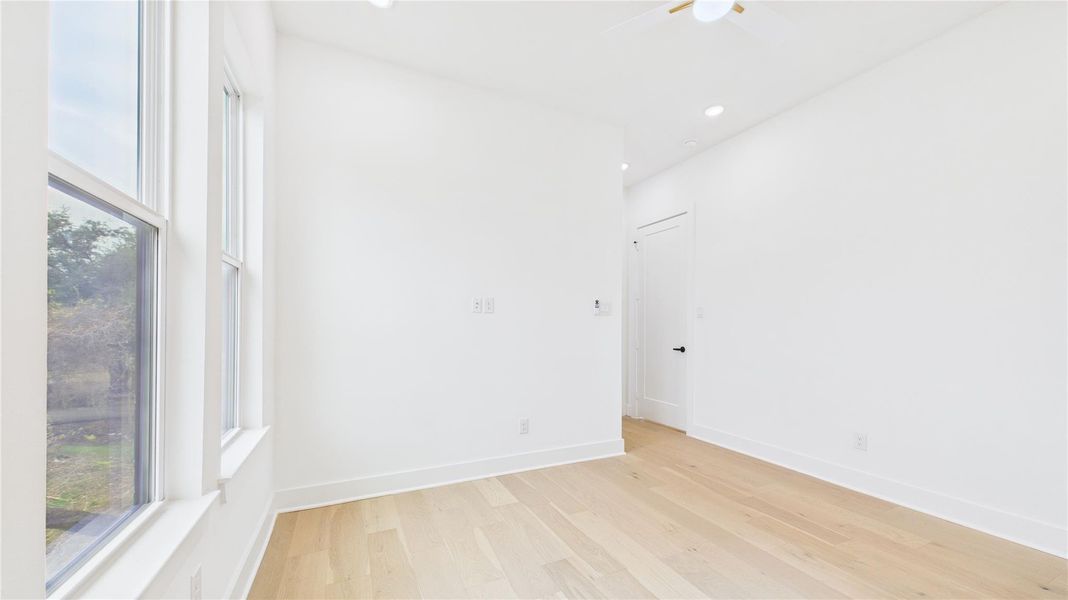 This room is bright and airy with large windows letting in natural light. It features light wood flooring and white walls, creating a clean, modern look. A ceiling fan is present, and there’s a door leading to another area.