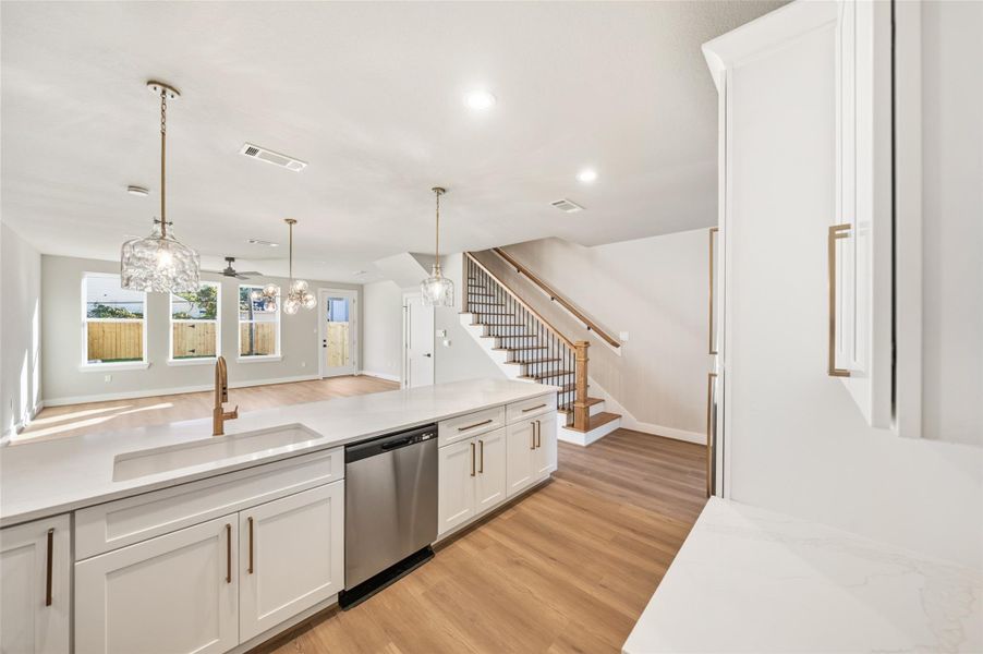 Open-concept kitchen and living area. It features a spacious island with a sink, elegant pendant lighting, and stainless steel appliances. The area has light wood flooring, ample natural light from multiple windows, and a staircase leading to the upper floor.