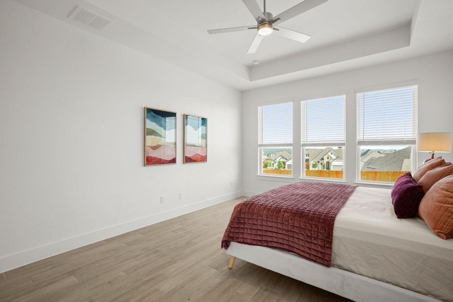 A well-lit bedroom features a tray ceiling, ceiling fan, and large windows offering neighborhood views.
