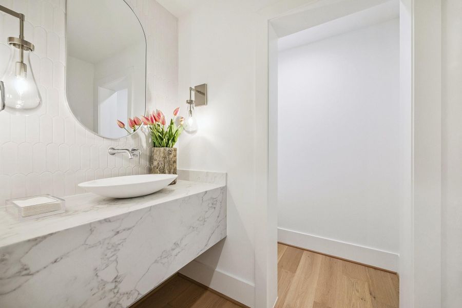 Second Floor Powder Room with a Floating Slab Quartz Counter, Oval Stone Composite White Vessel Sink, Wall-Mounted Widespread Faucet in Polished Chrome. The backsplash wall is adorned with White Scallop Porcelain Mosaic Tile. There is also a Oval Framed Mirror and Two Raindrop Wall Mounted Sconces in Chrome.