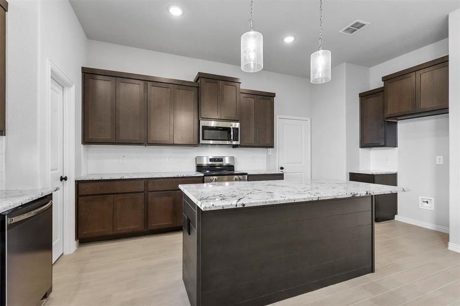 Kitchen featuring stainless steel appliances, dark brown cabinets, pendant lighting, light hardwood / wood-style floors, and light stone countertops