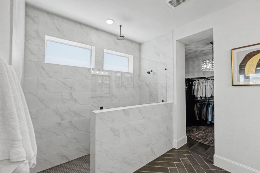 The extensive primary shower offers a luxurious rain showerhead and two bright transom windows set within sleek tile walls. A half-height tiled wall with a glass panel separates the wet area. This view leads into a substantial walk-in closet featuring a dark patterned floor runner, a crystal chandelier, and plenty of hanging space.