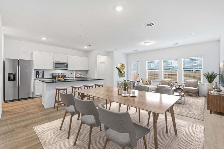 Open-concept living area featuring wood-finish flooring, a kitchen with white cabinetry and black countertops, stainless steel appliances, and multiple windows providing natural illumination