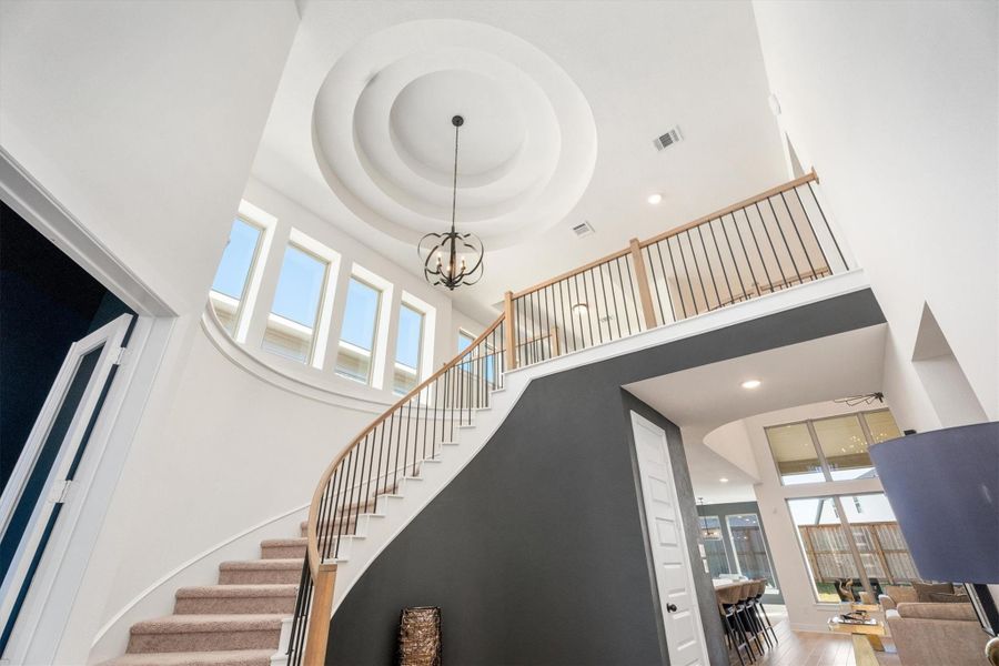 This photo showcases a spacious and elegant entryway featuring a grand curved staircase with wooden railings. The high ceiling includes an ornate light fixture, and large windows flood the area with natural light. The open-concept design leads into a bright living space with modern decor.
