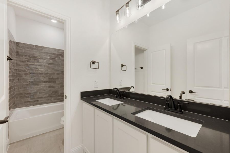 Bathroom with double sink vanity, black countertop, white cabinets, and tiled shower-tub combo visible through doorway