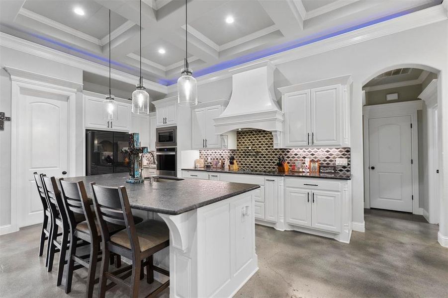 Kitchen featuring white cabinetry, beamed ceiling, decorative light fixtures, premium range hood, and a kitchen island with sink