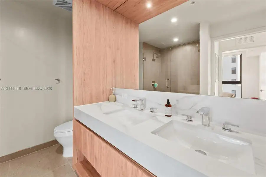 Double vanity featuring a light wood-finish cabinet, white solid-surface countertop with dual integrated sinks, polished chrome widespread faucets, and a full-width mirror