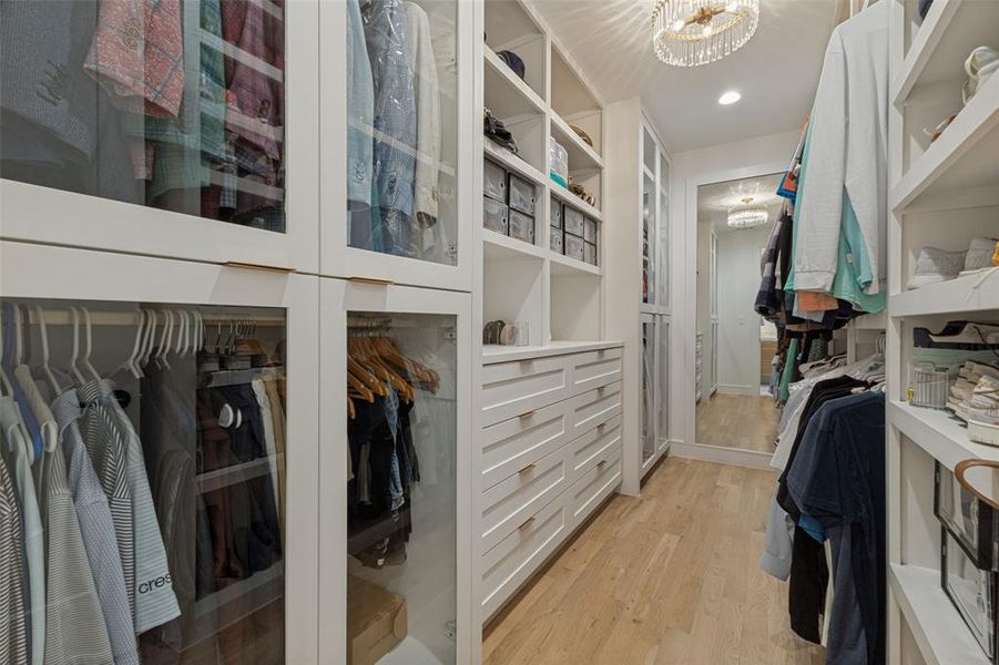 Custom closet system featuring white cabinetry, glass-front storage, built-in drawers, open shelving, and wood-finish flooring Custom closet system featuring white cabinetry, glass-front storage, built-in drawers, open shelving, and wood-finish flooring