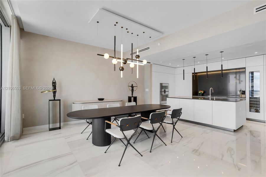 Open-concept interior featuring large-format stone flooring, a center island with integrated sink, built-in panel-ready appliances, and a geometric chandelier with linear and spherical lighting elements