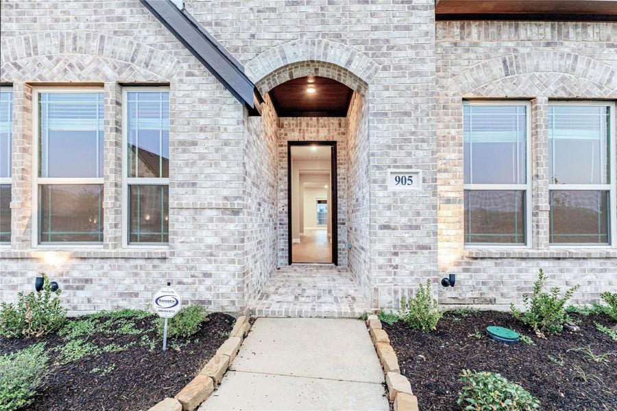 Brick home entrance with arched doorway, wood ceiling detail, and concrete walkway leading to front door