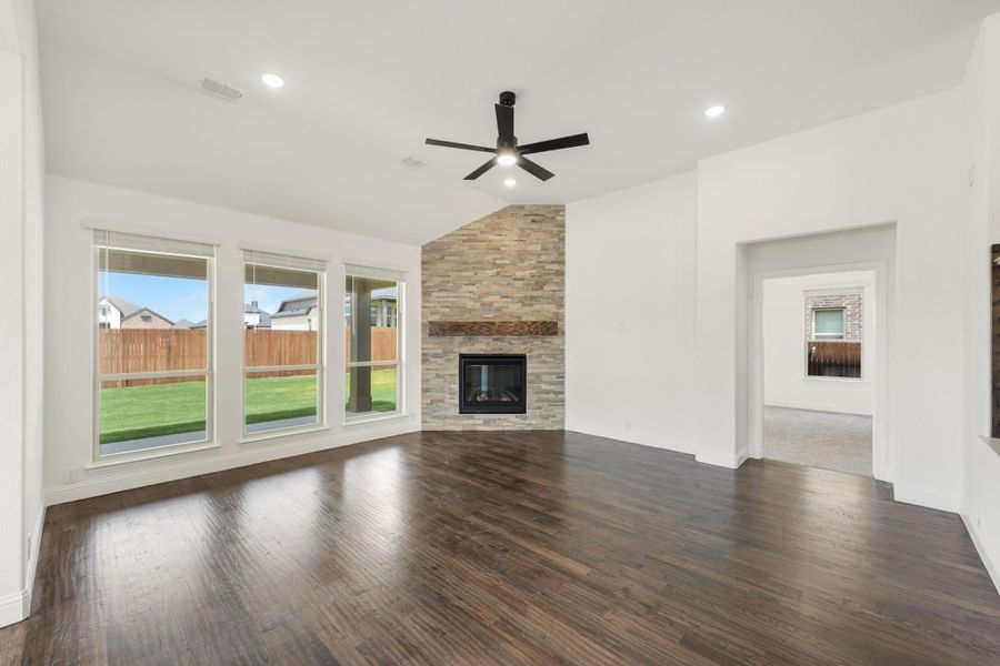 Open living room with dark hardwood floors, stone fireplace, ceiling fan, and large windows overlooking backyard