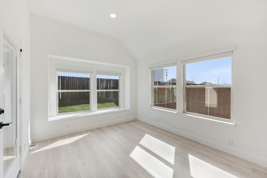 Empty bedroom with vaulted ceiling, light wood floors, and multiple windows overlooking backyard
