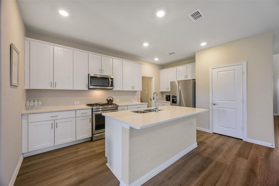 Modern kitchen featuring an island with an undermount sink, white cabinetry, stainless steel appliances, light-toned countertops, and wood-finish flooring Modern kitchen featuring an island with an undermount sink, white cabinetry, stainless steel appliances, light-toned countertops, and wood-finish flooring