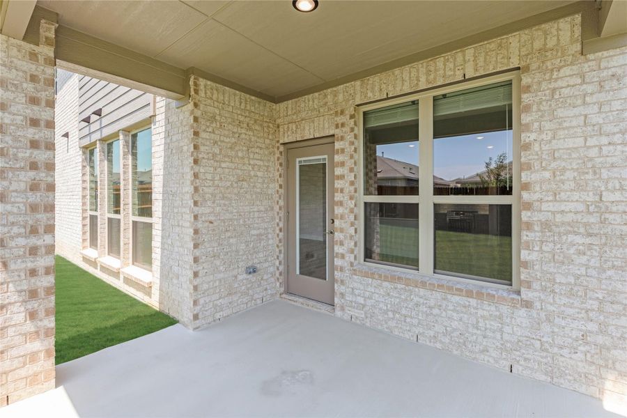 Covered back patio with concrete floor, light brick exterior, rear entry door, and large window overlooking green yard