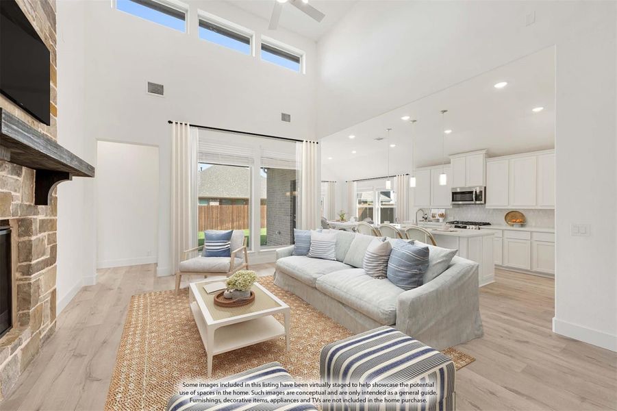 Open-concept living room with two-story ceiling, stone fireplace, and connected white kitchen with hardwood floors