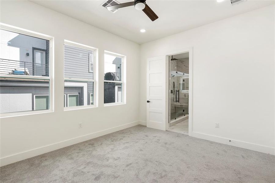 This is a bright, modern room with neutral walls and carpet, featuring three large windows that let in plenty of natural light and a ceiling fan for comfort. An en suite bathroom, adding convenience and style.