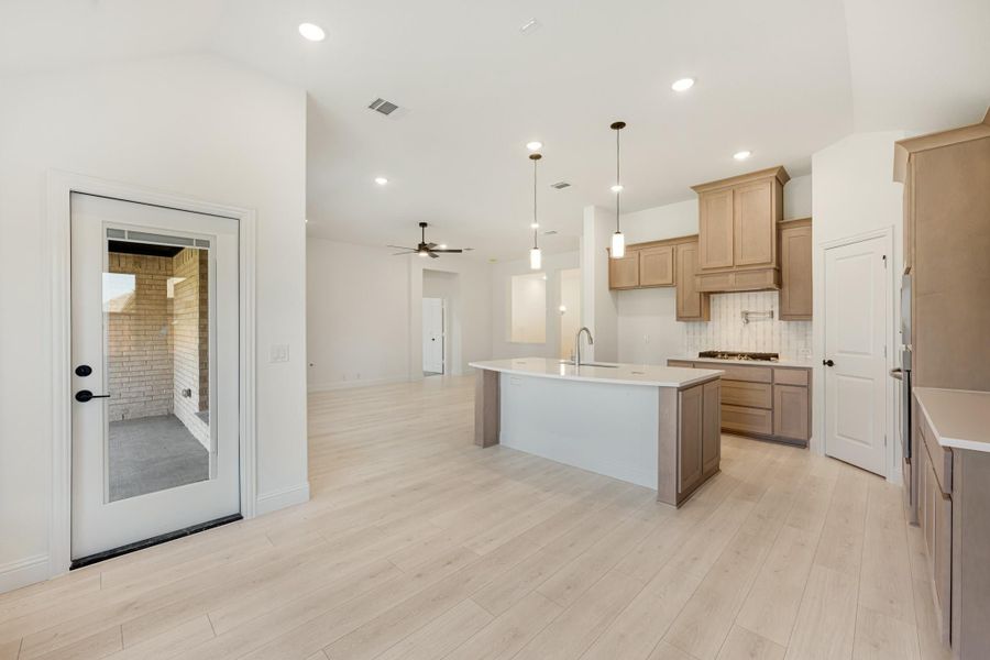Open-concept kitchen with center island, tan cabinets, pendant lights, and light wood floors leading to living area