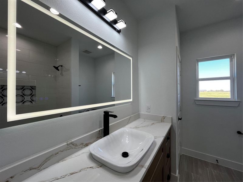 Modern bathroom with a sleek design featuring a large illuminated mirror, stylish black fixtures, and a vessel sink on a quartz countertop. The space is brightened by natural light from a window with a scenic view.