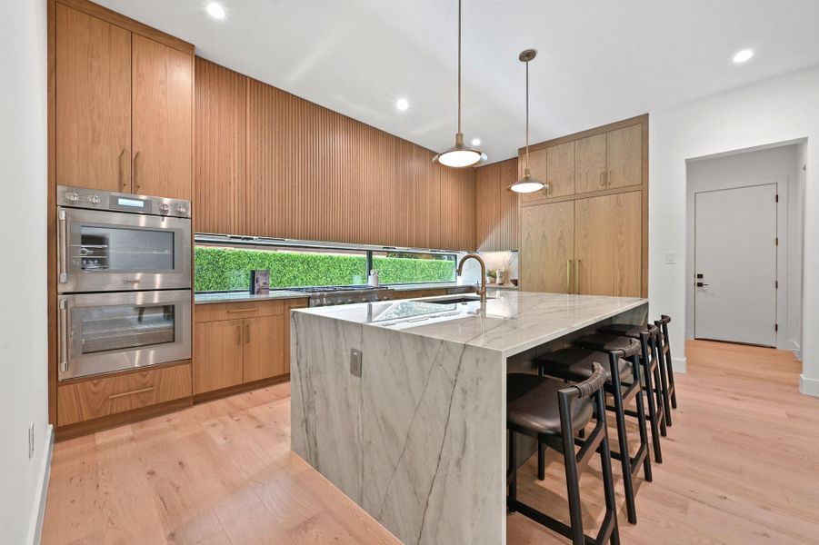 Kitchen featuring light stone countertops, a kitchen bar, double oven, light wood-style floors, and decorative light fixtures