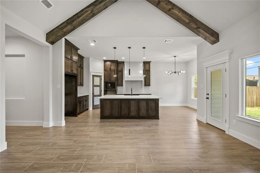 Open concept living space featuring a kitchen island with seating, vaulted ceiling with exposed wood beams, wood-look tile flooring, shaker-style cabinetry, and a glass-paneled exterior door