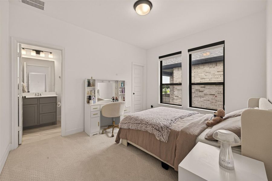 This photo shows a cozy bedroom with a neutral color palette, featuring a comfortable bed, a stylish vanity with a chair, and large windows offering natural light. The room connects to a bathroom with modern fixtures, enhancing convenience and privacy.