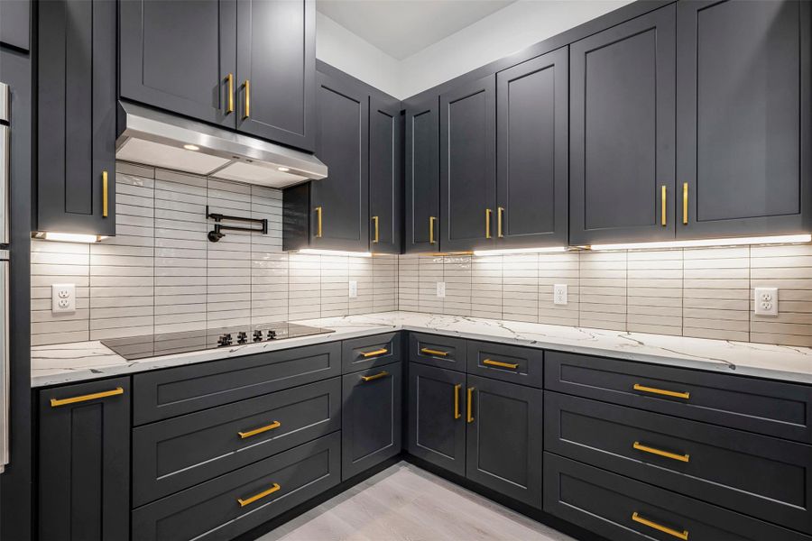 Kitchen featuring an under-cabinet range hood, light stone countertops, tasteful backsplash, black electric stovetop, and light wood-finished floors. Kitchen featuring an under-cabinet range hood, light stone countertops, tasteful backsplash, black electric stovetop, and light wood-finished floors.