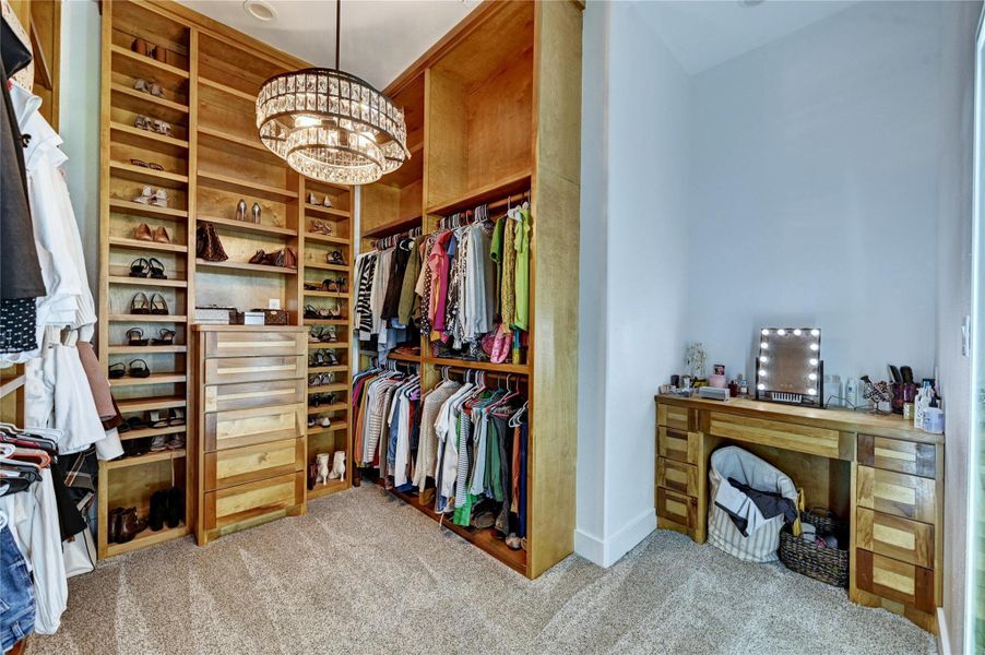 This spacious walk-in closet features custom wooden shelving, ample hanging space, and a luxurious chandelier. It includes a stylish vanity area with a mirror, perfect for dressing and organizing.