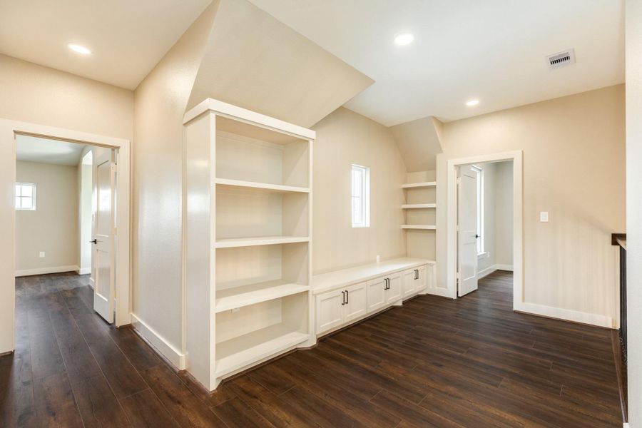 This photo showcases a bright, open space with built-in shelving and storage, dark hardwood floors, and neutral walls. It connects to adjacent rooms through doorways, providing a versatile area for a home office or reading nook.