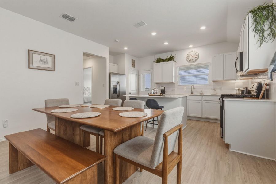 This open concept area features light flooring, a dining space with a wooden table, and a kitchen with white cabinetry and stainless steel appliances