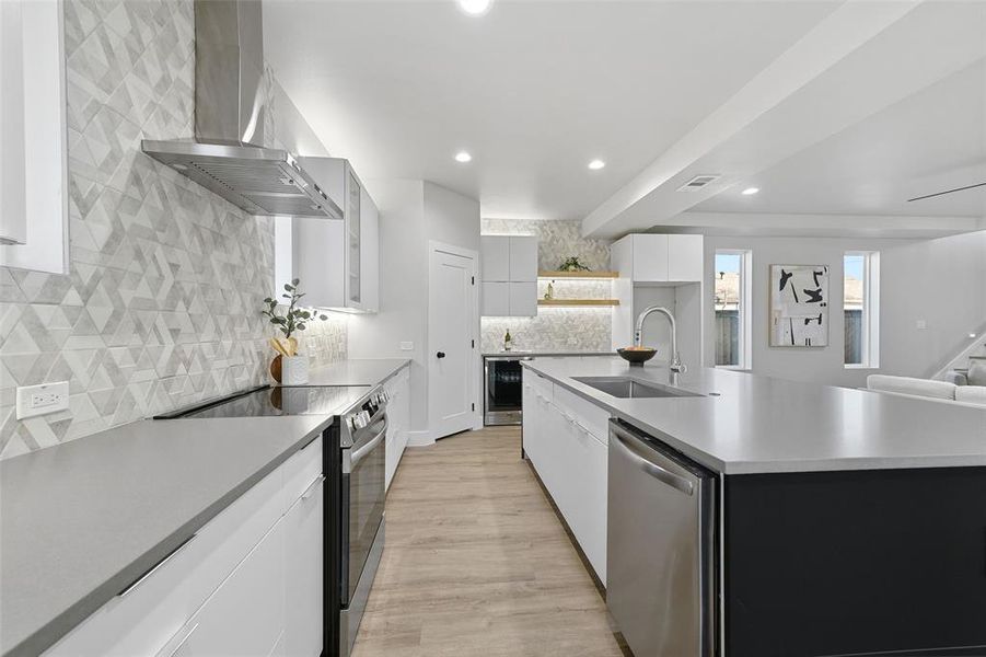 Kitchen with a large island with sink, sink, stainless steel appliances, white cabinets, and wall chimney exhaust hood