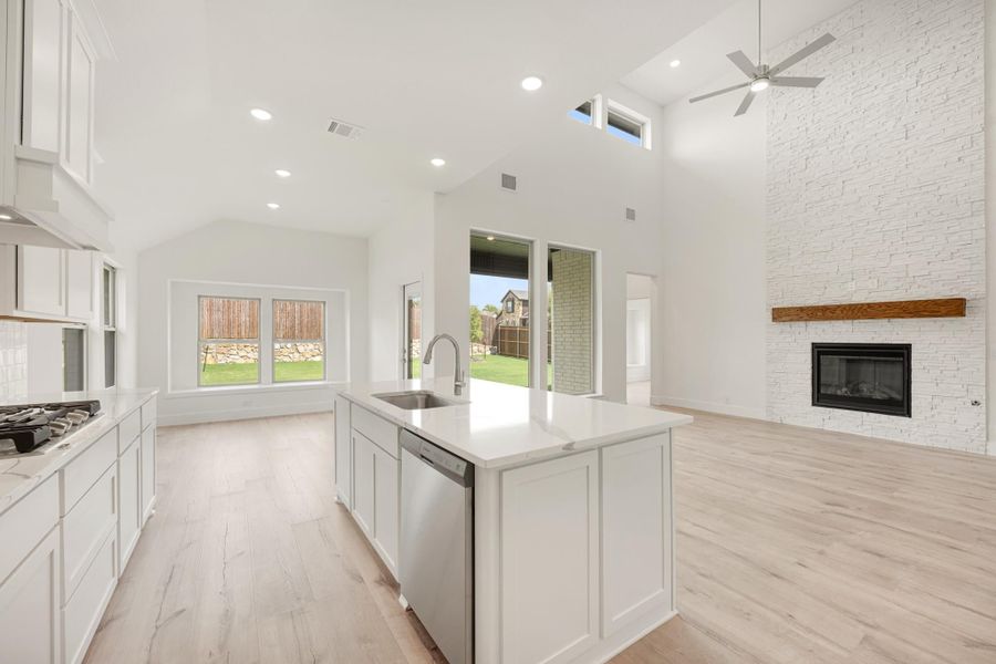 Open-concept kitchen with white island, quartz countertops, and stone fireplace with vaulted ceiling and wood floors