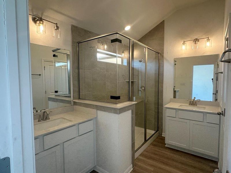 Bathroom featuring dark wood-style flooring, two vanities, a shower stall, vaulted ceiling, and a textured wall Bathroom featuring dark wood-style flooring, two vanities, a shower stall, vaulted ceiling, and a textured wall
