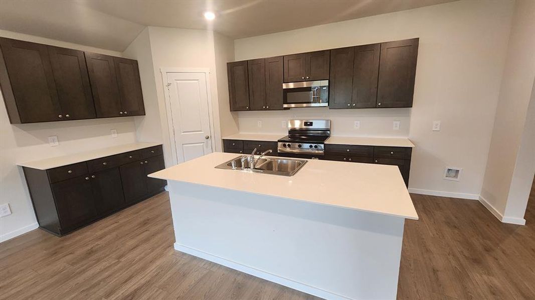 Kitchen featuring a center island with double basin sink, dark wood-finish cabinetry, light countertops, stainless steel appliances, and wood-finish flooring