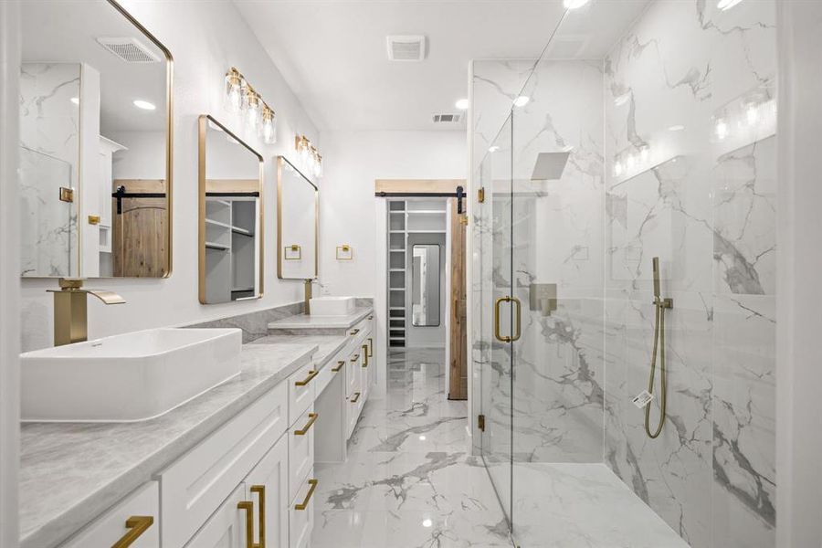 Spacious bath featuring a glass-enclosed shower with a rainfall showerhead, dual vanity with vessel sinks, white cabinetry, gold-finish hardware, and marble-look tile flooring