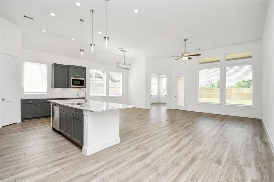 This is a bright, open-concept kitchen with modern  cabinetry and a central island, complemented by light wooden flooring. The space is well-lit with natural light from large windows and features contemporary pendant lighting.