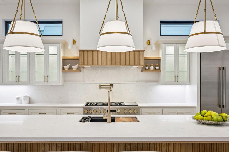 A closer view of this gorgeous kitchen showcases Slab Quartz Counter and Backsplash, 33 Inch Undermount Single Basin Sink in Quartz Composite Finish, Pull-Down Faucet in Champagne Bronze and Top Switch Disposal, 24“ Pot Filler in a Champagne Bronze Finish, White Oak Floating Shelves with Aged Gold/White Straight Swing Arm Pendant and LED Under-Cabinet Lighting and Under-Cabinet PlugMold.  Inset Cabinetry with Glass Fronted Doors and Shelves with Lighting on the Upper Cabinets.