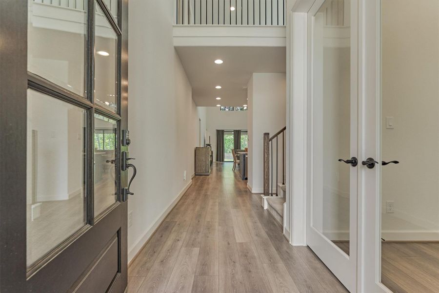 This photo showcases a modern, open-concept entryway with light wood flooring, high ceilings, and a view into a bright living space. There's a staircase on the right, and large windows at the back bring in natural light.