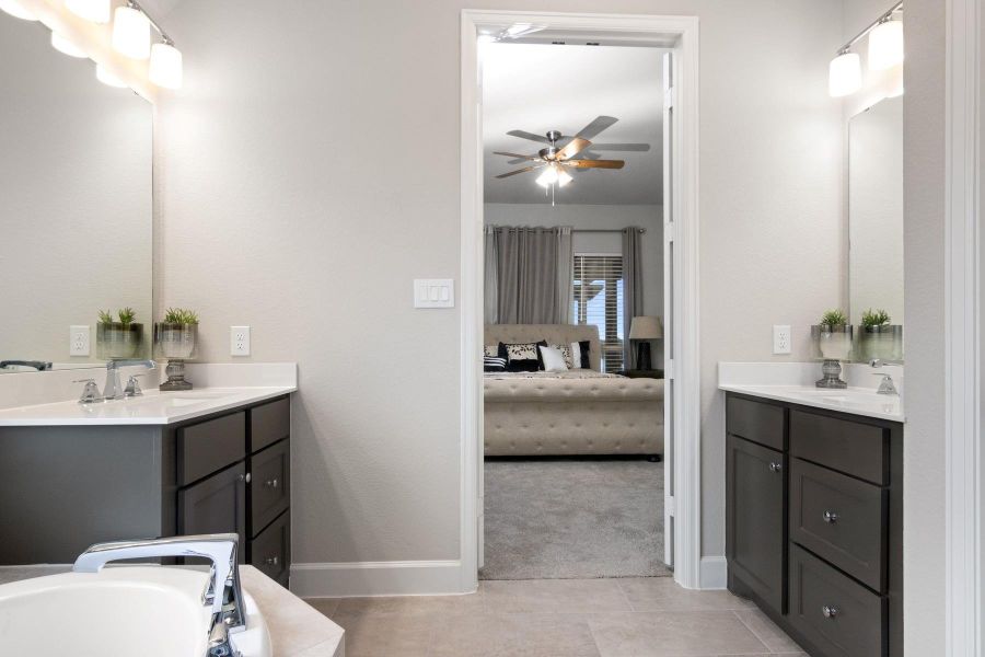 This photo showcases a modern bathroom with dual sinks, dark cabinetry, and elegant lighting. It opens into a cozy bedroom with a ceiling fan, and neutral decor, creating a seamless and inviting living space.