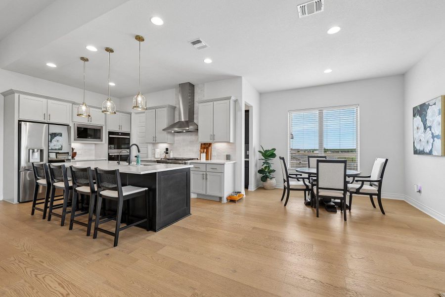 Modern kitchen featuring light gray cabinetry, stainless steel appliances, a dark-toned island with seating, pendant lighting, and wood-finish flooring