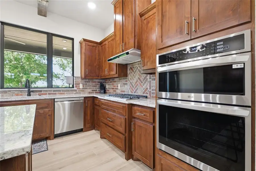 Kitchen featuring warm wood cabinetry, light granite countertops, stainless steel appliances, a herringbone brick-patterned backsplash, and wood-finish flooring